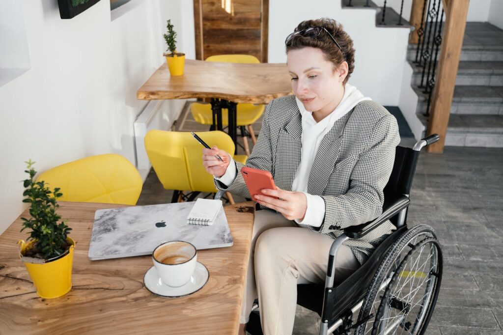 High angle of focused female in wheelchair browsing cellphone at table with netbook and cup of latte